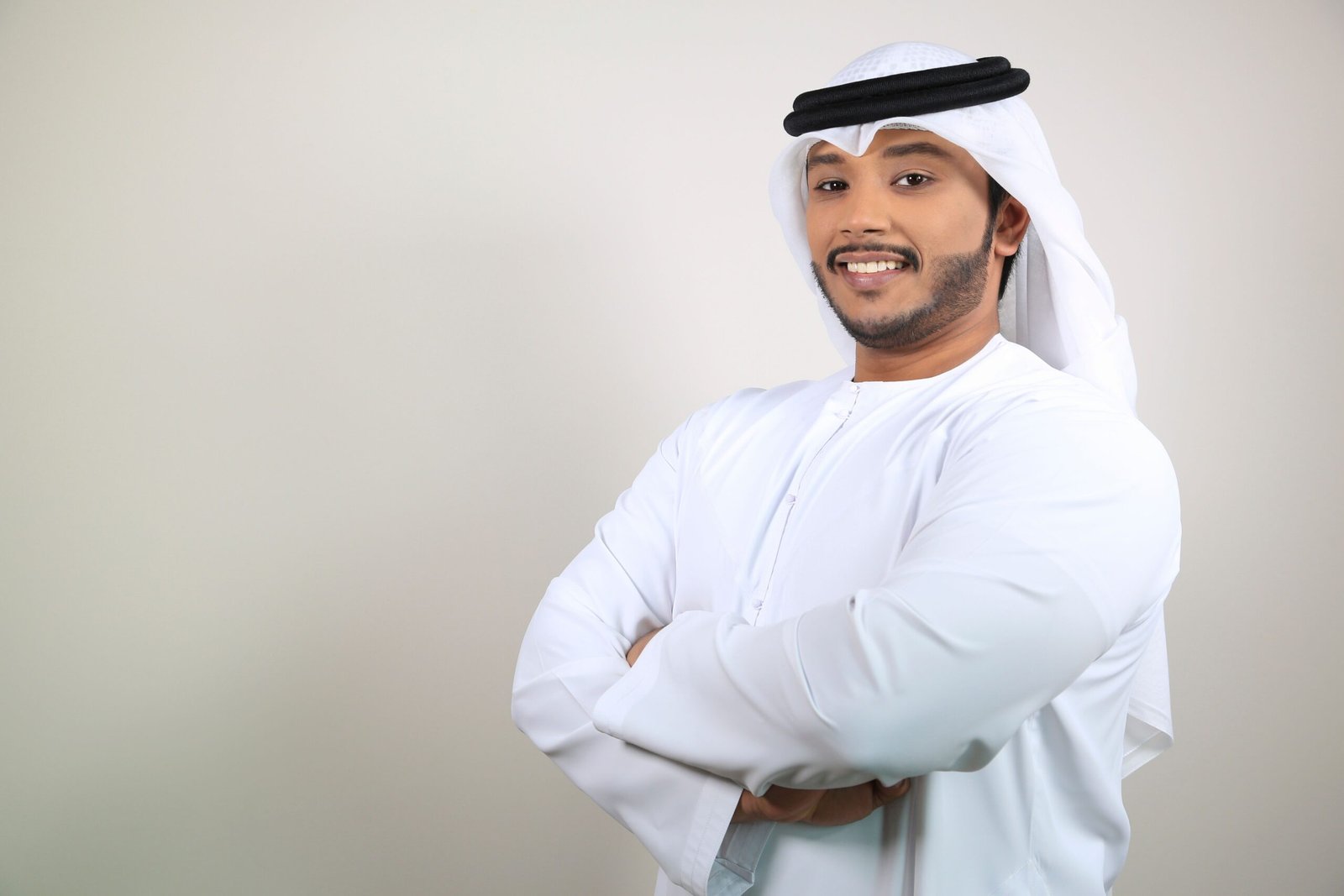 Smiling Middle Eastern man in a white kandura crossing his arms, exuding confidence indoors.